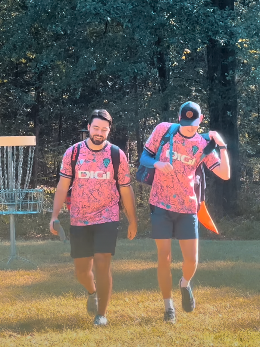 Two men wearing matching pink jerseys with a 'Digi' logo walk through a grassy area near a disc golf basket, with trees in the background.